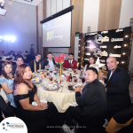 A group of people in formal attire sits around a table at a decorated event venue. A "10th Anniversary" sign is displayed on a screen in the background.