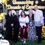 A group of five people, some wearing masks, stand in front of a banner reading "Unmasking a Decade of Excellence." One person in the center holds a certificate. Event sponsors' logos are visible at the bottom.