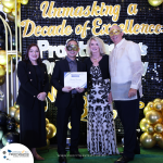 Four people stand together at an event, two wearing masquerade masks. One person holds a certificate. A backdrop with text "Unmasking a Decade of Excellence" and decorative balloons is in the background.