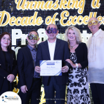 A group of people wearing masks poses for a photo at an event. One person in the center holds a certificate. The background reads "Unmasking a Decade of Excellence".