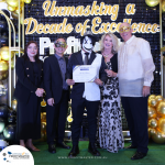 A group of five people, four adults and one youth, wearing formal attire and masquerade masks, stand in front of a backdrop reading "Unmasking a Decade of Excellence." The youth holds a certificate.