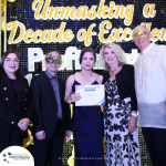 A group of five people dressed in formal attire stand together at an event with a banner reading "Unmasking a Decade of Excellence". One person in the center holds a certificate.