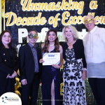 A group of five people in formal attire, some wearing masks, stand in front of a backdrop that reads "Unmasking a Decade of Excellence." The person in the center holds a certificate.