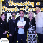 A group of five people at an event themed "Unmasking a Decade of Excellence." One person in the center is holding a certificate. Some individuals are wearing masks.