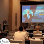 A man stands at a podium addressing an audience seated at tables, with a presentation slide overhead welcoming Richard and displaying his photo. The event is associated with Profitmaster Global Outsourcing.