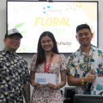 Three people in floral outfits standing in front of a "Floral Day!" event banner. The woman in the center holds an envelope.