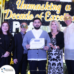 A group of five people, some wearing masks, pose for a photo. The person in the center holds a certificate. A sign in the background reads "Unmasking a Decade of Excellence.