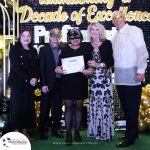 A group of five people, four wearing masks, stand together holding a certificate. They are dressed formally, with a backdrop stating "Decade of Excellence.