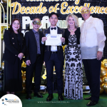 A group of five people, some wearing masks, stand together on a stage holding a certificate. A backdrop reads "Decade of Excellence." Balloon decorations surround the stage.