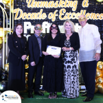 A group of five people, some wearing masks, pose for a photo during an event titled "Unmasking a Decade of Excellence." One person holds a certificate. Balloons and event branding are in the background.