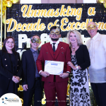 A group of six people wearing masquerade masks stand together. One man in the center, wearing a red suit, holds a certificate. Golden letters in the background read "Unmasking a Decade of Excellence.