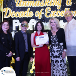 Five people dressed formally, some wearing masks, pose for a photo at an event. A woman in a red dress holds a certificate. A sign in the background reads "Unmasking a Decade of Excellence.