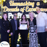 A group of five people, some wearing masquerade masks, stand in front of a decorated backdrop. The center person holds a certificate.