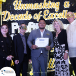 A group of five people poses for a photo at an event with a backdrop labeled "Unmasking a Decade of Excellence." One person in the center holds a certificate. Some individuals wear decorative masks.