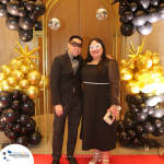 A man and woman wearing masks stand in front of a festive balloon arch with gold star decorations. They pose on a red carpet with a banner that reads "www.profitmaster.com.au" at the bottom.