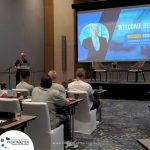 A speaker addresses an audience in a conference room with a presentation slide displaying "Welcome Reception" and an image of Richard Crookes, Founder and President of Profitmaster Global Outsourcing.