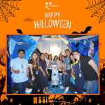 A group of people in a Halloween-themed setting, standing around a person in a snowman costume. The backdrop features Halloween decorations with the text "Happy Halloween.