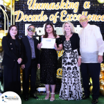 A group of five people, three wearing masks, stand on a stage. A woman in the center holds a certificate. The backdrop reads, "Unmasking a Decade of Excellence.