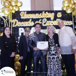 A group of five people, some wearing masks, pose for a photo in front of a sign that reads "Unmasking a Decade of Excellence." One man in the center holds a framed certificate.