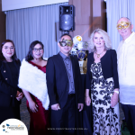 A group of five people in formal attire and masks pose for a photo at an event. A banner and a decorative trophy are in the background.