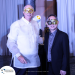 Two men wearing decorative masks pose together at an event. The backdrop features white curtains and a sign that reads "PROFITMASTER." One man is in a barong tagalog, the other in a dark suit.