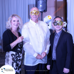 Three people stand together at an event, each wearing ornate masks. They are dressed formally, with one person in traditional attire. The backdrop features drapes and lighting fixtures.