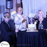 Three people in masquerade masks stand behind a black and gold cake on a table, celebrating Profitmaster's 50th anniversary. A logo and website URL are visible at the bottom of the image.