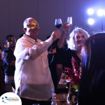 Two people wearing masks raise their wine glasses in celebration at an indoor event with dim lighting. Other attendees and decorations are visible in the background.