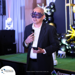 A man in a suit and dark mask speaks into a microphone while holding a phone. He stands on a decorated stage with balloons and lights in the background.