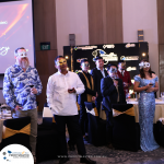 A group of people wearing formal attire and decorative masks stand in a room with tables and chairs. A banner in the background displays logos and the text "Profitmaster.