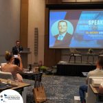 A man stands at a podium giving a presentation, while a projected slide behind him displays his name, photo, and the topic of his talk on anti-money laundering. An audience is seated in front of him.