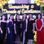 A group of people dressed in formal attire stands in front of a decorated backdrop reading "Unmasking a Decade of Excellence," with balloons and lights adding to the celebratory atmosphere.