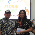 Three people wearing floral shirts stand in front of a floral-themed sign.