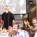 People wearing decorative masks are celebrating an anniversary at an indoor event. Some are seated and clapping, while one person is standing. A large screen in the background displays "Anniversary.