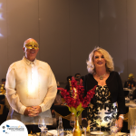 A man wearing a traditional outfit and a gold mask stands next to a woman in a black and white dress at a formal event. The table in front of them is decorated with a vase of red flowers.