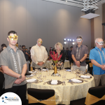 A group of people stands around a banquet table, wearing masks. The table is set with dishes, glasses, and a flower centerpiece. A banner in the background reads "Profitmaster.