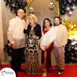 Four people wearing masquerade masks pose for a photo at a formal event, standing on a red carpet with a balloon arch in the background.