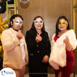 Three people wearing masquerade masks pose indoors. They make hand gestures and are dressed in formal attire. Balloons and a banner with "Profit Master" branding are in the background.