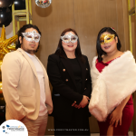 Three individuals wearing formal attire and masquerade masks stand together in front of a decorated background with gold star balloons. A sign on the bottom reads "Profitmaster Group.