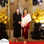 Two women in formal attire and decorative masks stand beneath an archway decorated with black and gold balloons at a themed event. A sign on the floor displays a logo and website URL.