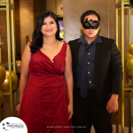 A woman in a red dress and a man in a suit with a black mask stand together at an event. The backdrop includes balloons.