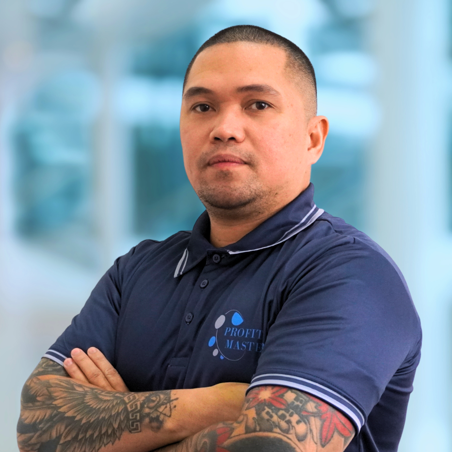 A bald man with tattoos on his arms stands with his arms crossed, wearing a blue polo shirt bearing the "Profit Masters" logo. Representing Our Team, he exudes confidence in the blurred background.