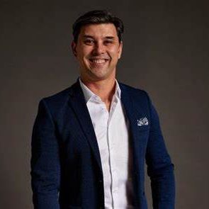 Anton Thysee, wearing a dark blue blazer, white shirt, and pocket square, smiles against a plain dark background.