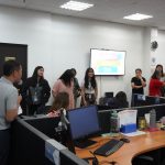 A group of people stands and talks near desks in an office. A digital screen displays a presentation titled "Maker's Day.