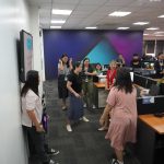 A group of people standing in an office, engaged in conversation and laughter near desks and computer monitors.