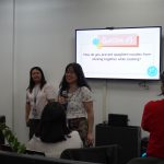 A woman holds a microphone, standing near a screen displaying "Question #5: How do you prevent spaghetti noodles from sticking together while cooking?" Other people are in the room listening.