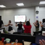 People in an office room are standing and clapping. A person at the center is holding a microphone near a screen displaying text. Desks with computers are visible in the foreground.