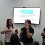 People in a room singing with microphones; a screen displays lyrics in Tagalog in the background.