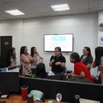 Group of people standing and sitting in an office, some holding microphones. They are gathered around a presentation screen displaying text.