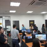 A group of people stands in an office room with computers, listening attentively. Some are seated while others are standing around a central workspace.